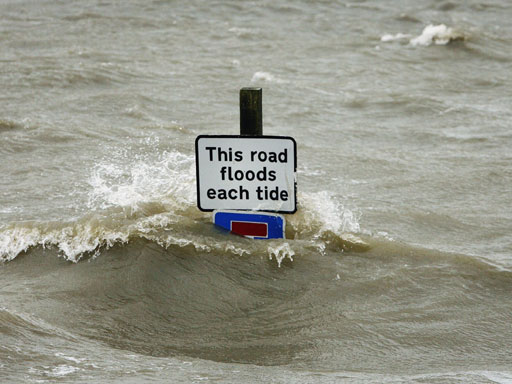 A traffic information sign with the text "This road floods each tide". The sign is partially submerged by coastal flooding.