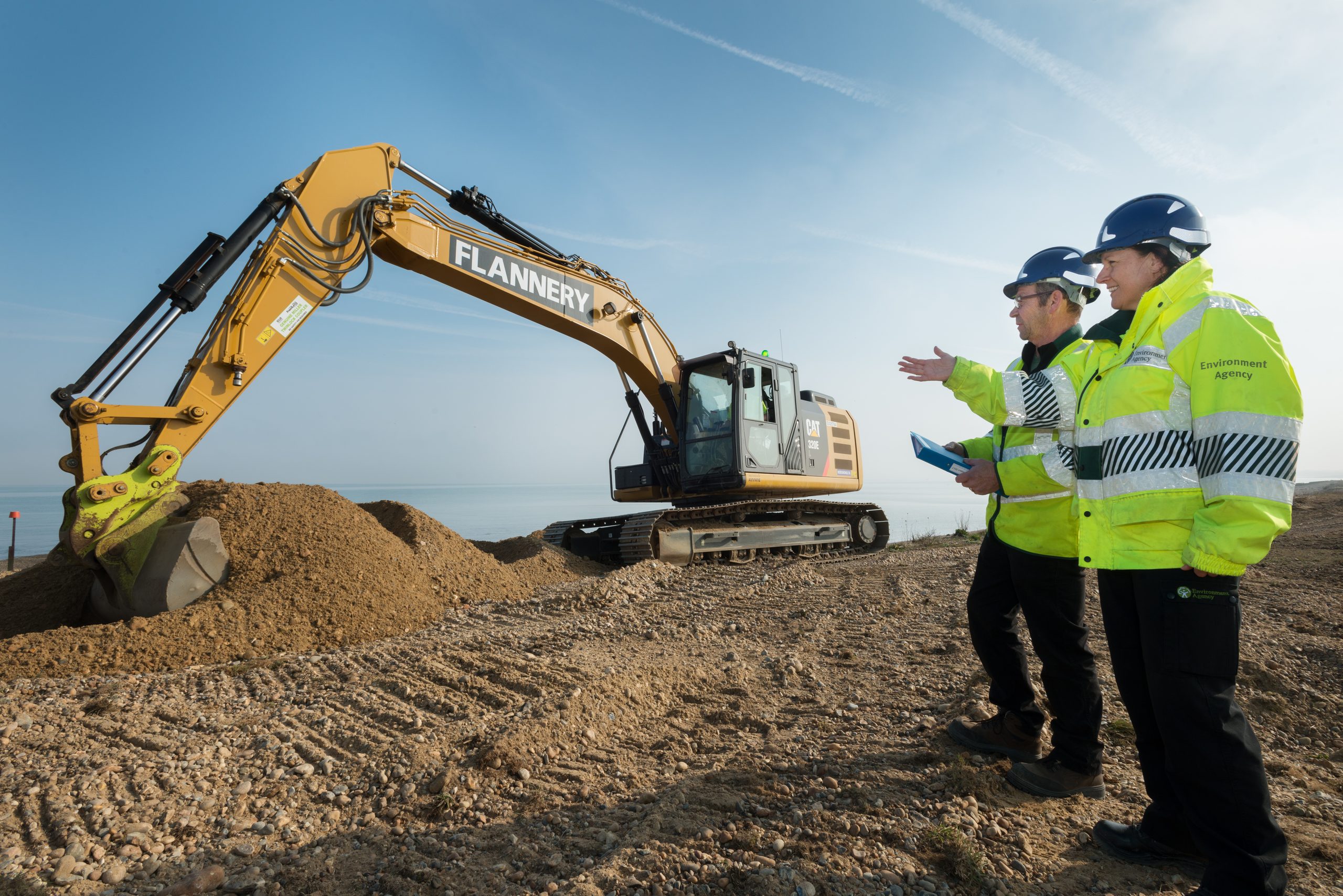 Two Environment Agency staff stand beside a digger carrying out shingle repairs.