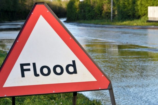 A traffic warning sign with the text "Flood". The sign is next to a flooded road.